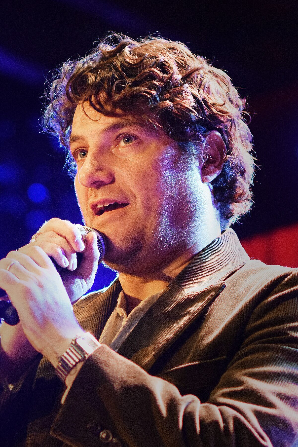 A photo of Adam Pally holding a microphone and speaking into it.